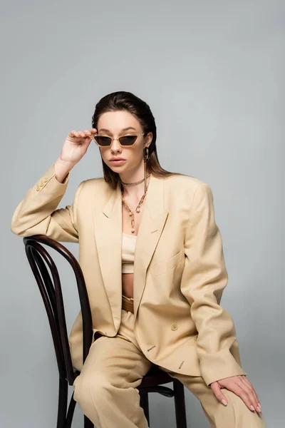 Young woman in stylish beige outfit and trendy sunglasses sitting on wooden chair isolated on grey - foto de stock