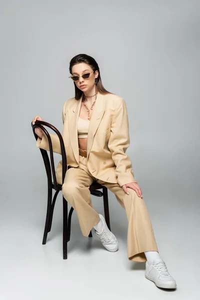 Full length of young woman in stylish beige outfit and trendy sunglasses sitting on wooden chair on grey - foto de stock