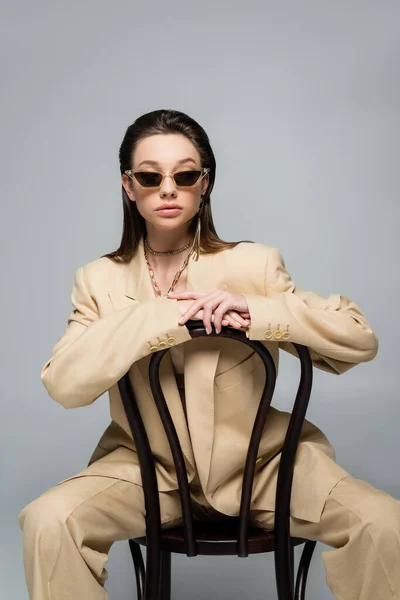 Brunette woman in stylish beige outfit and trendy sunglasses sitting on wooden chair isolated on grey — Fotografia de Stock