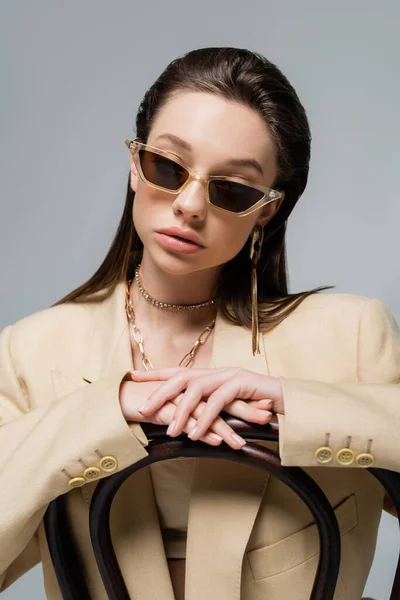 Young woman in stylish outfit and trendy sunglasses sitting on wooden chair isolated on grey — Stockfoto