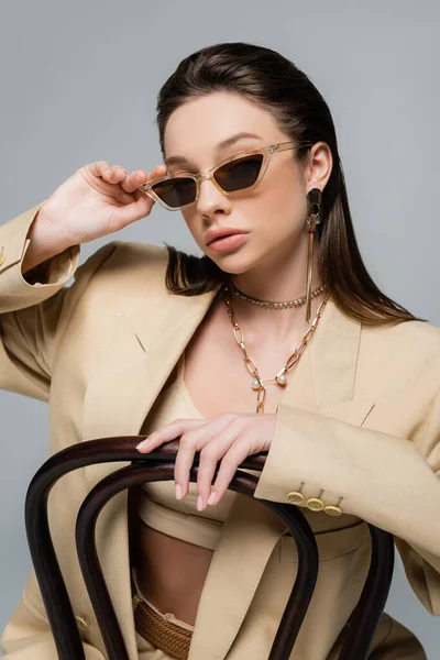 Young woman in stylish beige outfit adjusting trendy sunglasses while sitting on wooden chair isolated on grey - foto de stock