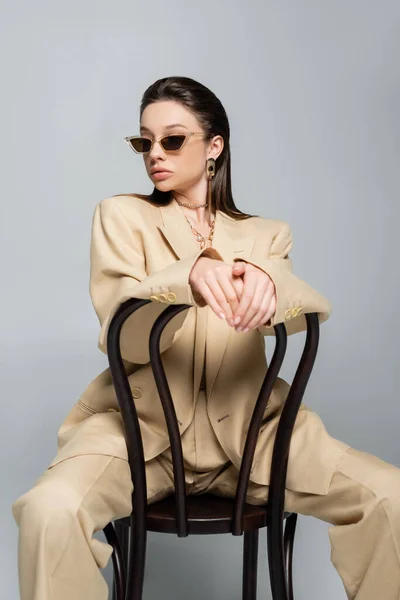 Молода жінка в стильному бежевому вбранні і сонцезахисних окулярах, що сидять на дерев'яному стільці ізольовані на сірому — стокове фото