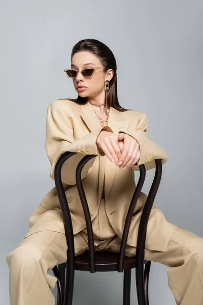 Young woman in stylish beige outfit and sunglasses sitting on wooden chair isolated on grey — Stock Photo