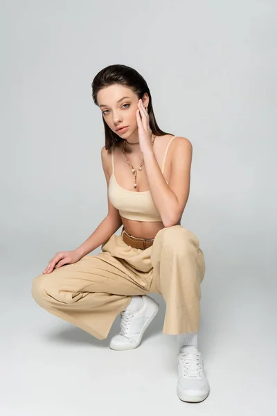 Full length of brunette woman in stylish beige outfit and trendy accessories posing while sitting on grey — Stock Photo