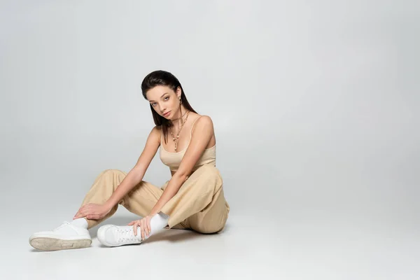 Full length of brunette woman in stylish beige outfit and trendy accessories sitting on grey — стоковое фото