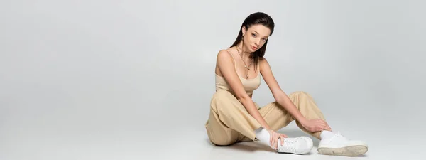 Full length of brunette woman in stylish beige outfit and trendy accessories sitting on grey, banner — Foto stock