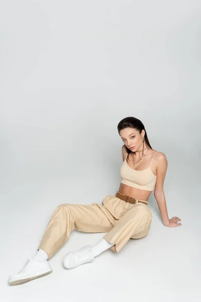 High angle view of brunette woman in stylish beige outfit and trendy accessories sitting on grey — стоковое фото