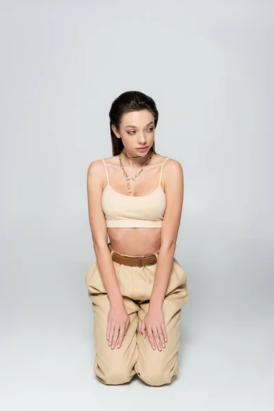 Brunette woman in stylish beige outfit and trendy accessories standing on knees on grey — Photo de stock