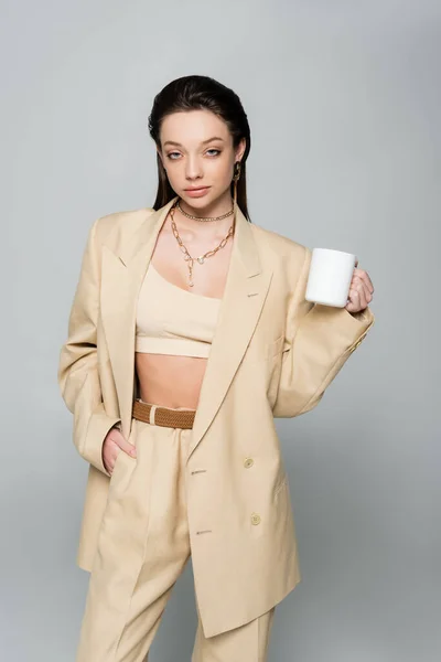 Brunette woman in stylish beige suit holding cup of coffee and posing with hand in pocket isolated on grey — Stock Photo