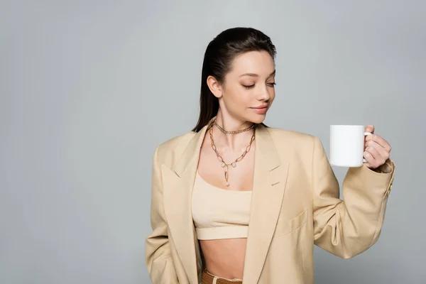 Cheerful woman in stylish beige suit holding cup of coffee isolated on grey — Foto stock