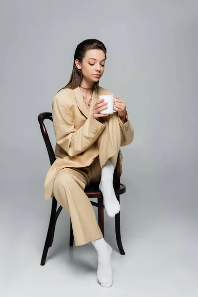 Full length of brunette woman in stylish beige suit holding cup of coffee and sitting on wooden chair on grey — Stockfoto