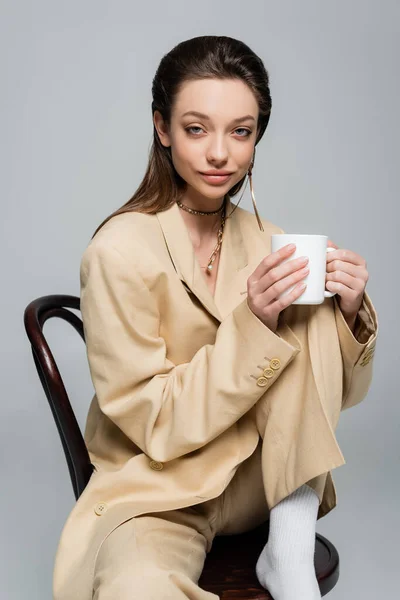 Брюнетка в стильному бежевому костюмі тримає чашку кави і сидить на дерев'яному стільці ізольовано на сірому — стокове фото