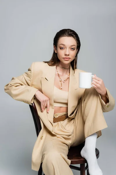 Pretty woman in stylish beige suit holding cup of coffee and sitting on wooden chair isolated on grey — Stockfoto