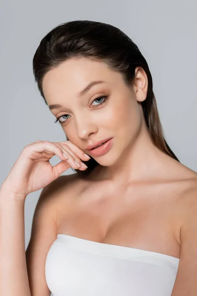 Portrait of smiling young woman with bare shoulders and makeup looking at camera isolated on grey - foto de stock