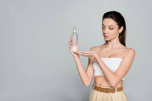 Young woman with bare shoulders holding bottle with cosmetic product isolated on grey — Stockfoto