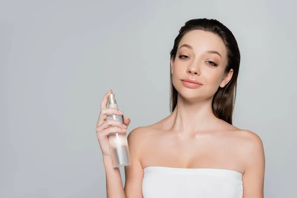 Young woman with bare shoulders holding bottle with face mist isolated on grey — Stock Photo