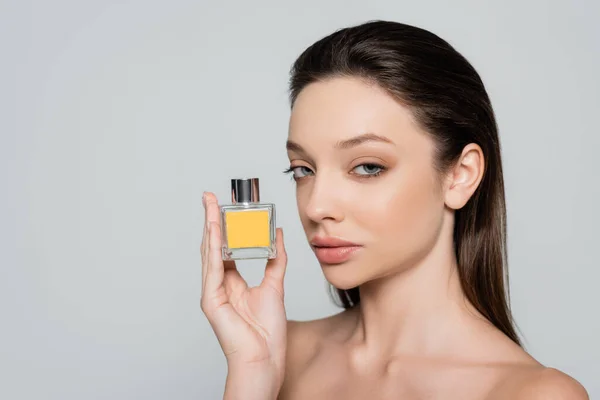 Pretty young woman with bare shoulders holding bottle with perfume isolated on grey — стоковое фото