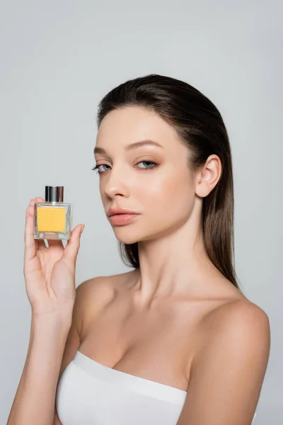 Pretty woman with bare shoulders holding bottle with luxury perfume isolated on grey - foto de stock