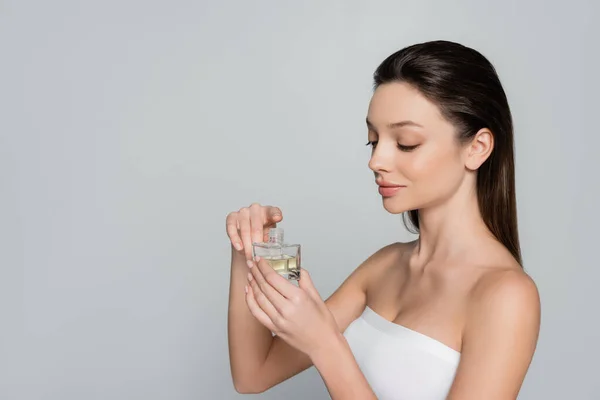 Young woman with naked shoulders holding bottle with luxury perfume isolated on grey — стоковое фото