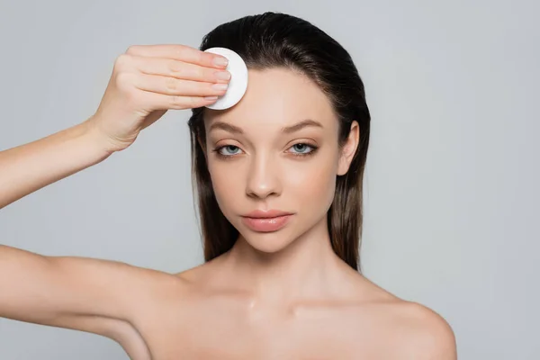 Young woman removing makeup on forehead with soft cotton pad isolated on grey — стоковое фото