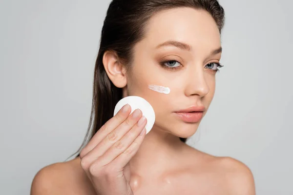 Young woman with bare shoulders and cosmetic product on face holding cotton pad isolated on grey — стоковое фото