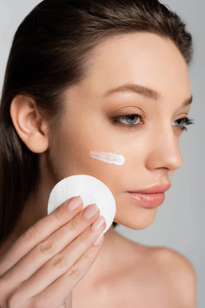 Close up of young woman with cosmetic product on face holding cotton pad isolated on grey — Stock Photo