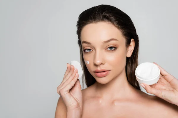 Young woman with bare shoulders and cream on face holding container and cotton pad isolated on grey — стоковое фото