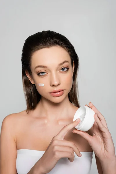 Brunette woman with bare shoulders and cream on face holding container isolated on grey — Photo de stock