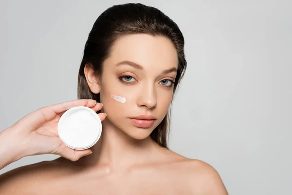 Young woman with bare shoulders and cream on face holding container isolated on grey — Stockfoto