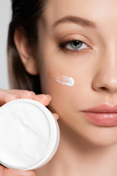 Close up view of cropped young woman with cosmetic cream on cheek holding container isolated on grey — Stock Photo