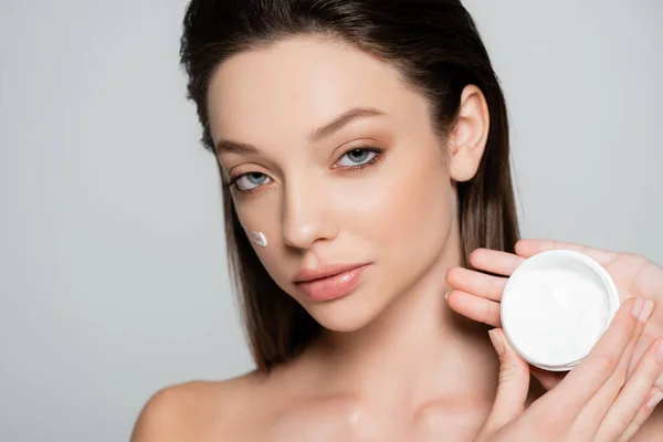 Brunette woman with bare shoulders and cosmetic cream on cheeks holding container isolated on grey — Foto stock