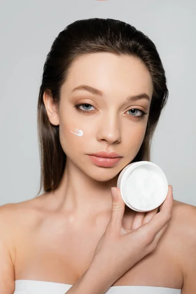 Young woman with cream on cheeks holding container isolated on grey — Photo de stock