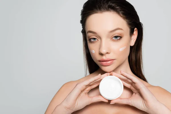 Young woman with bare shoulders and cream on cheeks holding container isolated on grey — Photo de stock