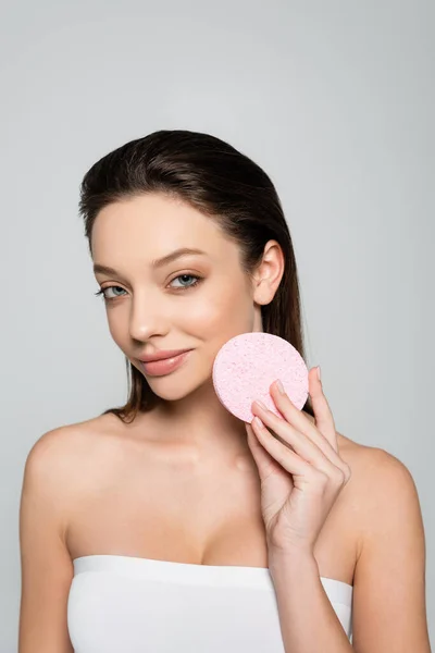 Smiling young woman with bare shoulders holding exfoliating sponge isolated on grey - foto de stock