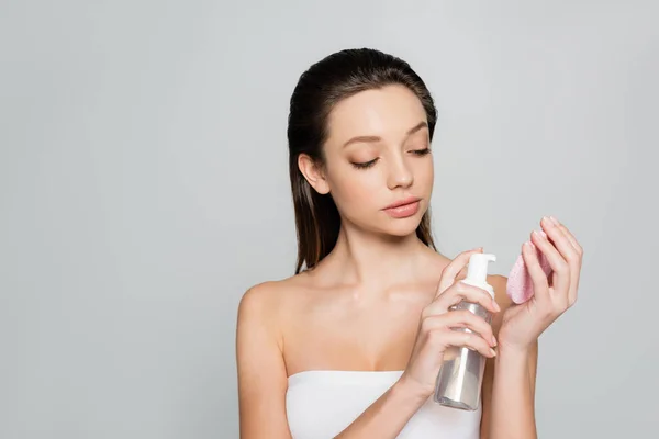 Young woman with bare shoulders holding bottle with cosmetic product near exfoliating sponge isolated on grey — Foto stock