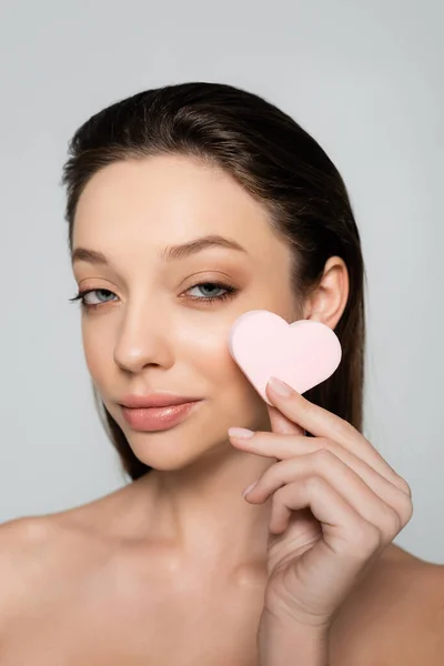 Young woman holding heart-shaped sponge near face isolated on grey — стоковое фото