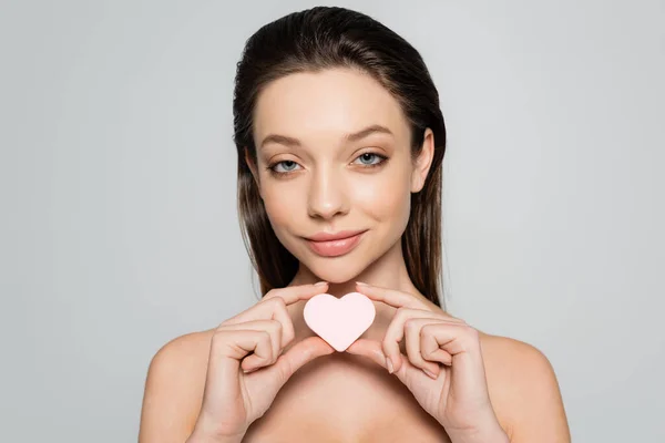 Smiling young woman holding heart-shaped sponge near face isolated on grey — Stockfoto