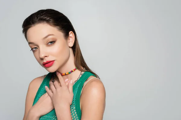 Young and stylish woman with red lips touching necklaces and posing isolated on grey — стоковое фото