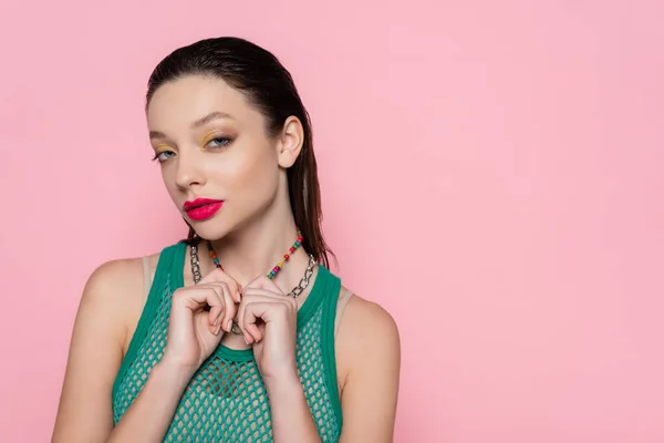 Young brunette woman with bright makeup pulling necklaces while looking at camera isolated on pink — Stockfoto