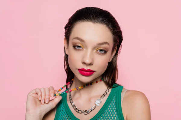 Young brunette woman with bright makeup pulling beads necklace while looking at camera isolated on pink — стоковое фото