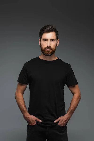 Portrait of bearded man in black t-shirt looking at camera while posing with hands in pockets isolated on grey — Photo de stock