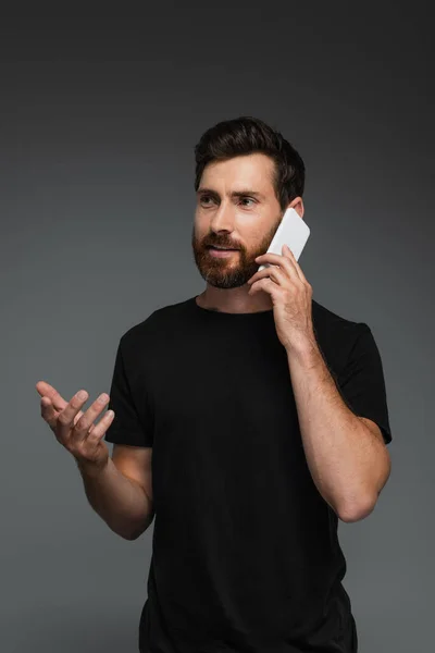 Бородатый мужчина в черной футболке разговаривает на смартфоне изолирован от серого — стоковое фото