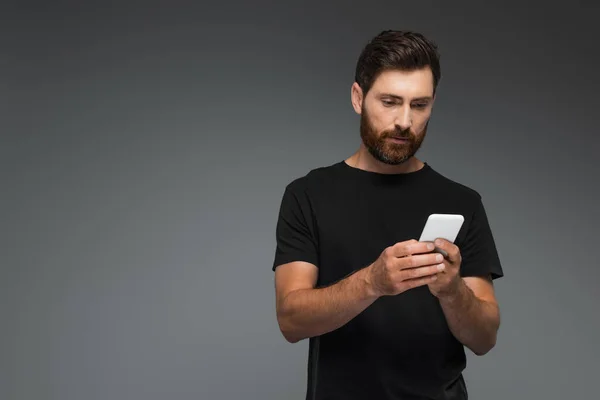 Bearded man in black t-shirt using smartphone isolated on grey — Stock Photo