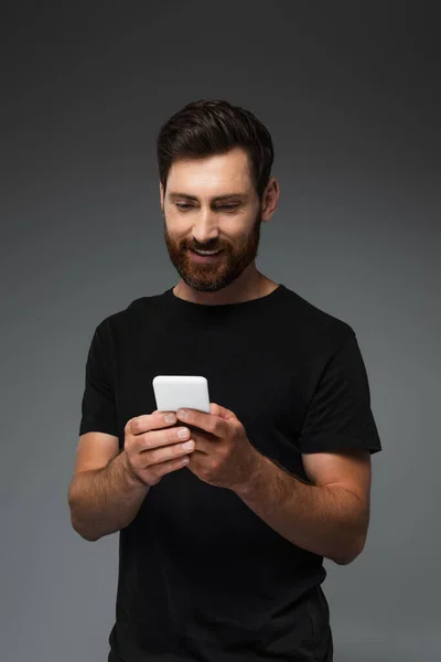 Happy bearded man in black t-shirt messaging on smartphone isolated on grey — Stock Photo