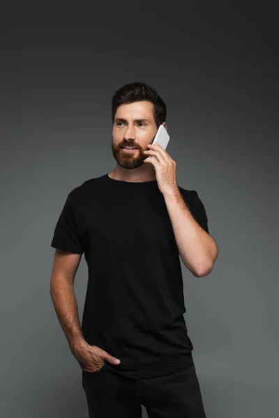 Bearded man in black t-shirt talking on smartphone while standing with hand in pocket isolated on grey — Stock Photo