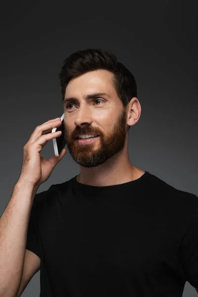Joyful and bearded man in black t-shirt talking on smartphone isolated on grey — Foto stock