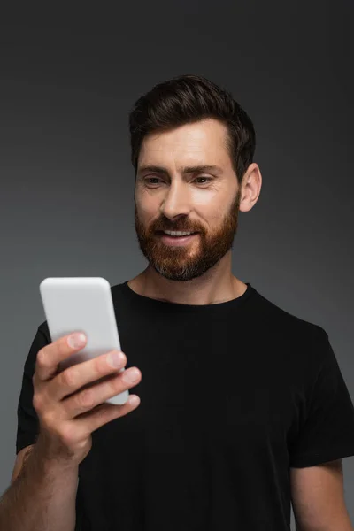 Pleased and bearded man in black t-shirt using mobile phone isolated on grey — Foto stock