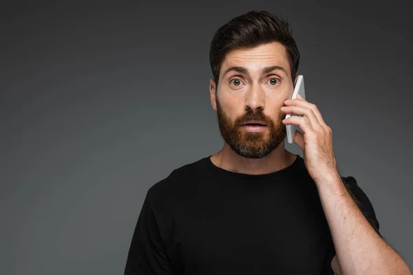 Shocked man in black t-shirt talking on smartphone isolated on grey — Stockfoto