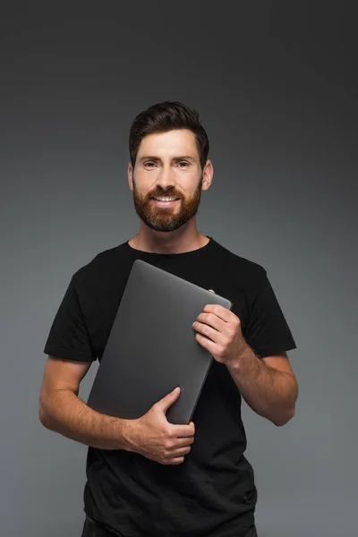 Щасливий бородатий чоловік у чорній футболці тримає ноутбук ізольований на сірому — стокове фото