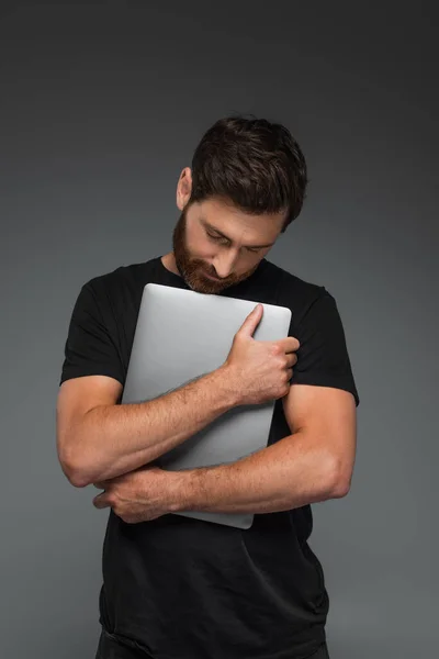 Бородатий чоловік в чорній футболці обіймає ноутбук ізольовано на сірому — Stock Photo